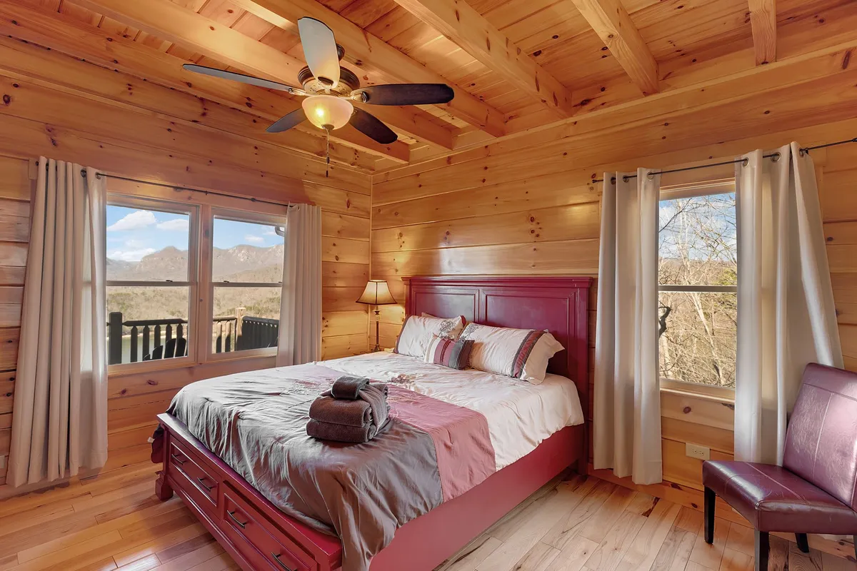 A cozy bedroom featuring a bed with a red headboard, large windows revealing a mountain view, and wood-paneled walls and ceiling. Soft natural light fills the space, highlighting the serene setting ideal for relaxation and comfort.