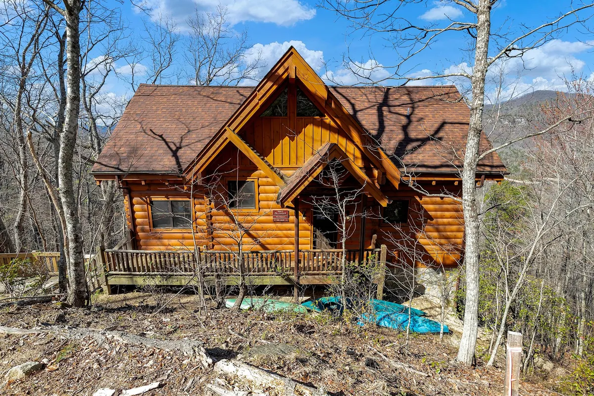 This image showcases the log cabin's exterior, featuring a welcoming front porch surrounded by lush trees and a stunning mountain view in the background. The inviting porch is set up for relaxation and socializing, connecting guests to the natural beauty around the property.
