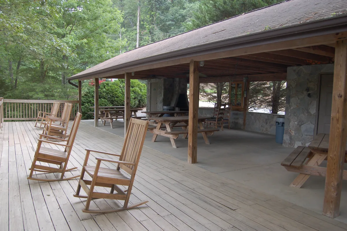 An inviting exterior shot with rocking chairs, picnic tables, wooden beams, and lush greenery framing the scene. The space is ideal for outdoor relaxation, morning coffee, or evening gatherings, blending privacy with the community feel.