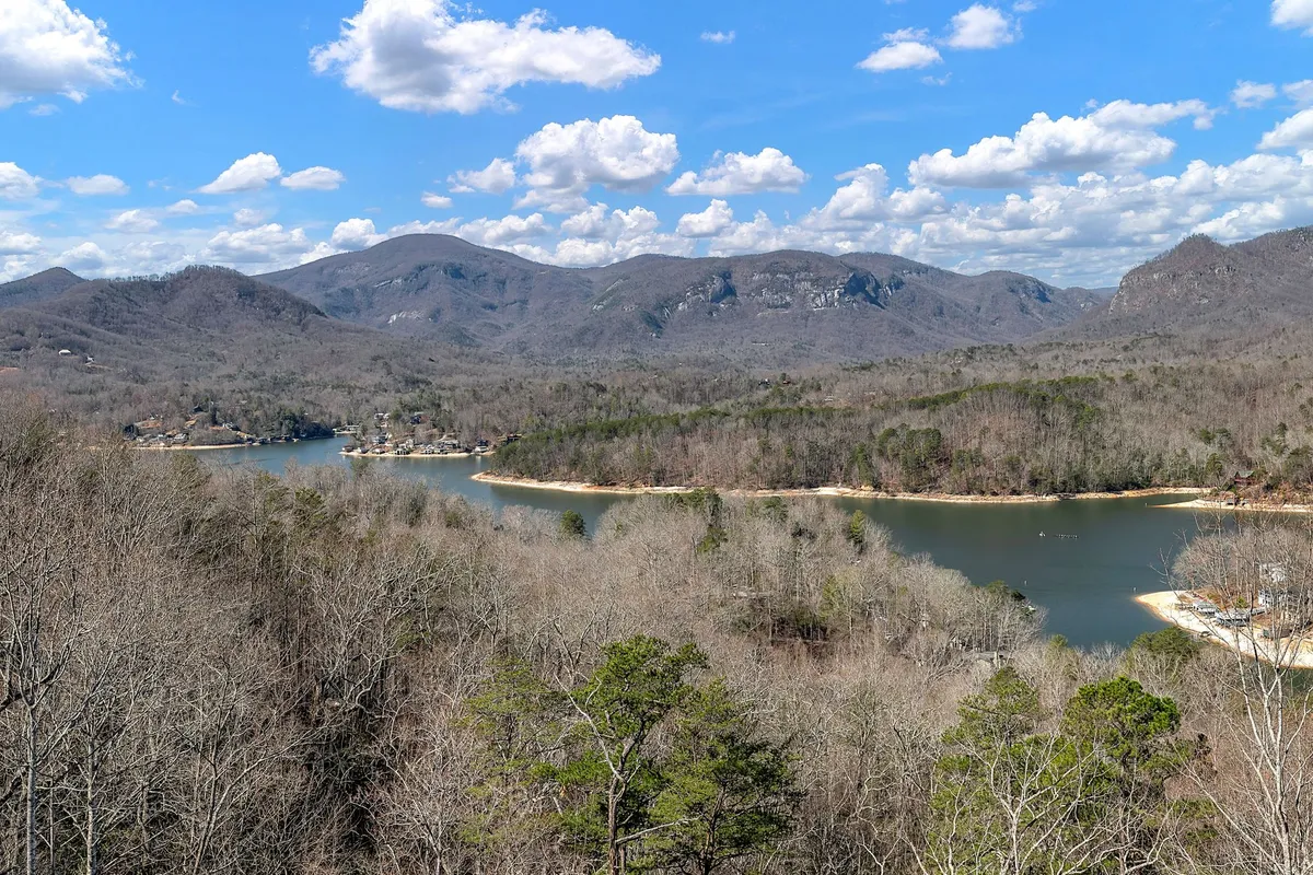 A sweeping view of a tranquil lake bordered by lush trees and set against the dramatic backdrop of distant mountains under a cloudy sky. The scene radiates serenity and natural beauty, inviting outdoor activities such as hiking, fishing, boating, and picnicking.