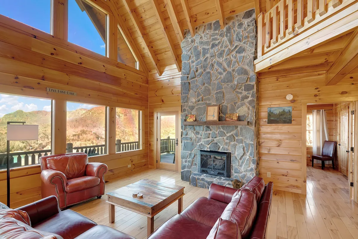 A cozy living room corner with a stone fireplace, leather sofas, wooden beams, and expansive windows letting in golden light and mountain scenery. The setting invites people to relax, share stories, or simply bask in the cabin’s warmth.