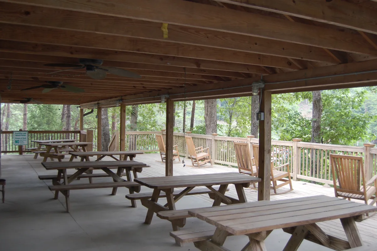 A relaxed outdoor gathering space with wooden picnic tables and rocking chairs overlooks a serene forested backdrop. The spacious porch is perfect for al fresco dining, group games, or simply soaking in the tranquil surroundings.