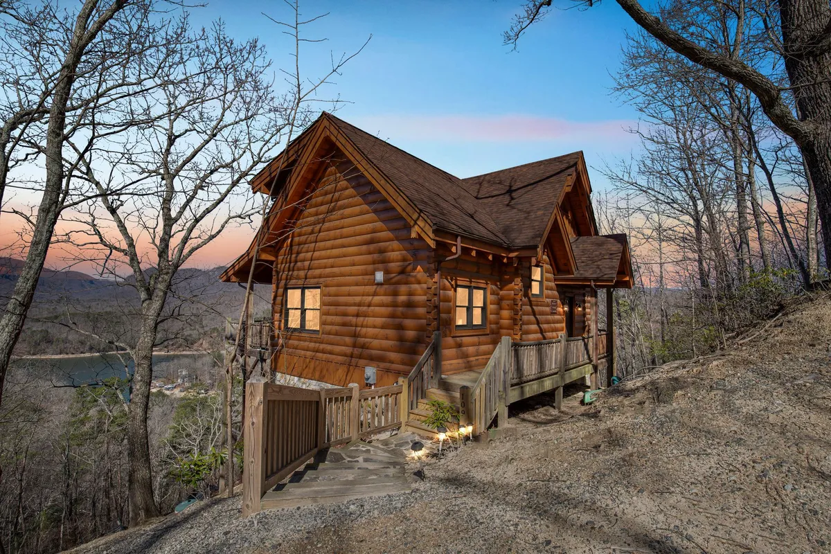 A log cabin nestled among tall trees, with a pathway leading to its welcoming entrance and distant mountain views beyond. The environment is inviting and serene, emphasizing a close connection to the outdoors and opportunities for hiking and nature exploration right from your doorstep.