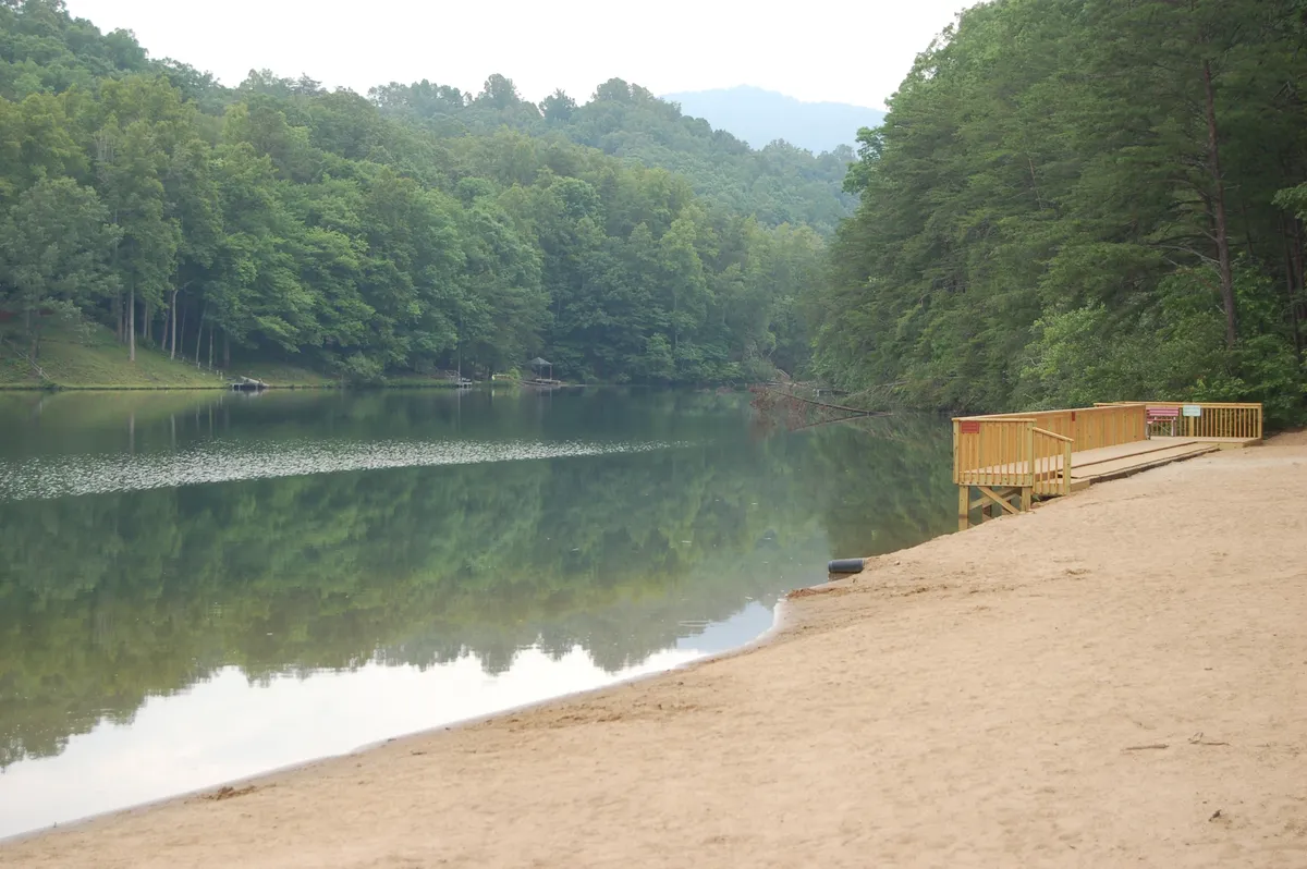 A tranquil scene featuring calm lake waters, a wooden dock, sandy beach, and surrounding trees. The setting invites activities like kayaking, swimming, and lakeside relaxation, with the private dock and beach foregrounded.