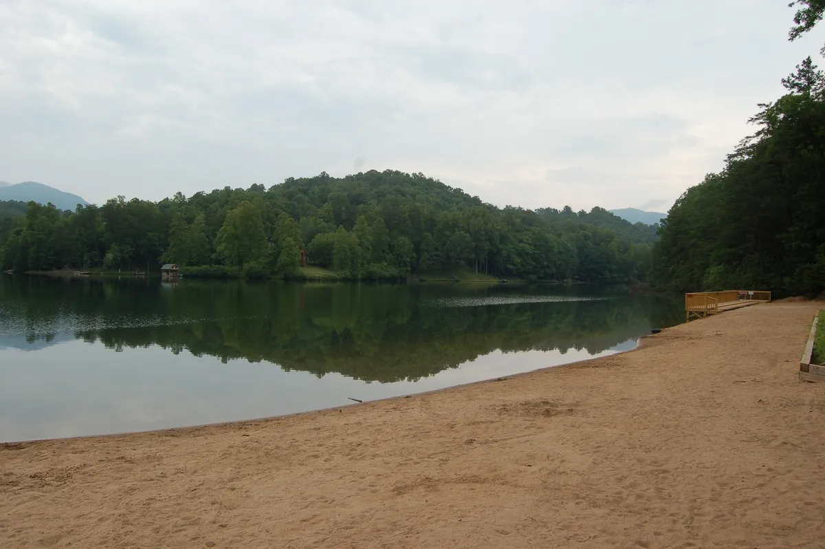 A peaceful lakeshore scene with calm water, green hills, and a sandy beach, set beneath a soft, golden sunset. The area is ideal for swimming, fishing, or simply relaxing and enjoying the beauty of the private waterfront.