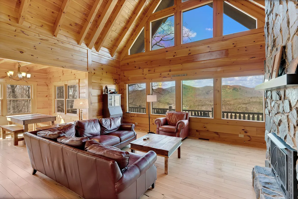 A sun-drenched open living area featuring large windows framing breathtaking mountain views. The space includes a prominent stone fireplace, plush leather sofas, and a wooden dining table set for a group, all tied together with warm, inviting colors and natural materials.