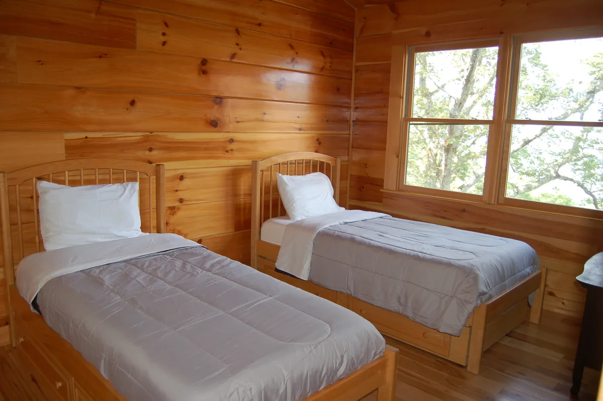 A bright, tranquil room with twin beds, wood-paneled walls, and expansive windows inviting in natural light and views. The space is simple yet comfortable, ideal for children or guests.