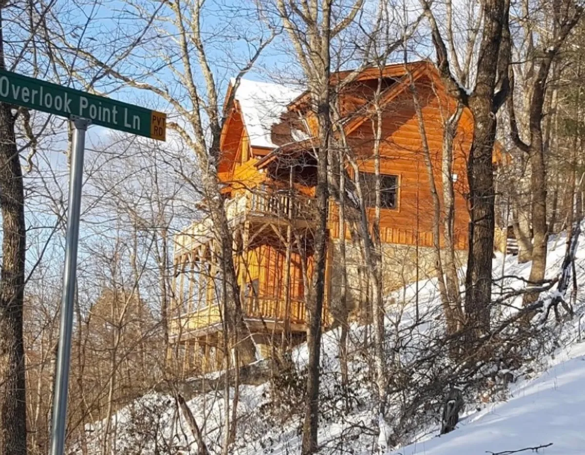 A log cabin nestled in a snow-covered landscape, with the Overlook Point Ln sign visible. The image evokes a sense of retreat and adventure, highlighting year-round appeal with opportunities for winter hiking, snowshoeing, and cozy evenings.