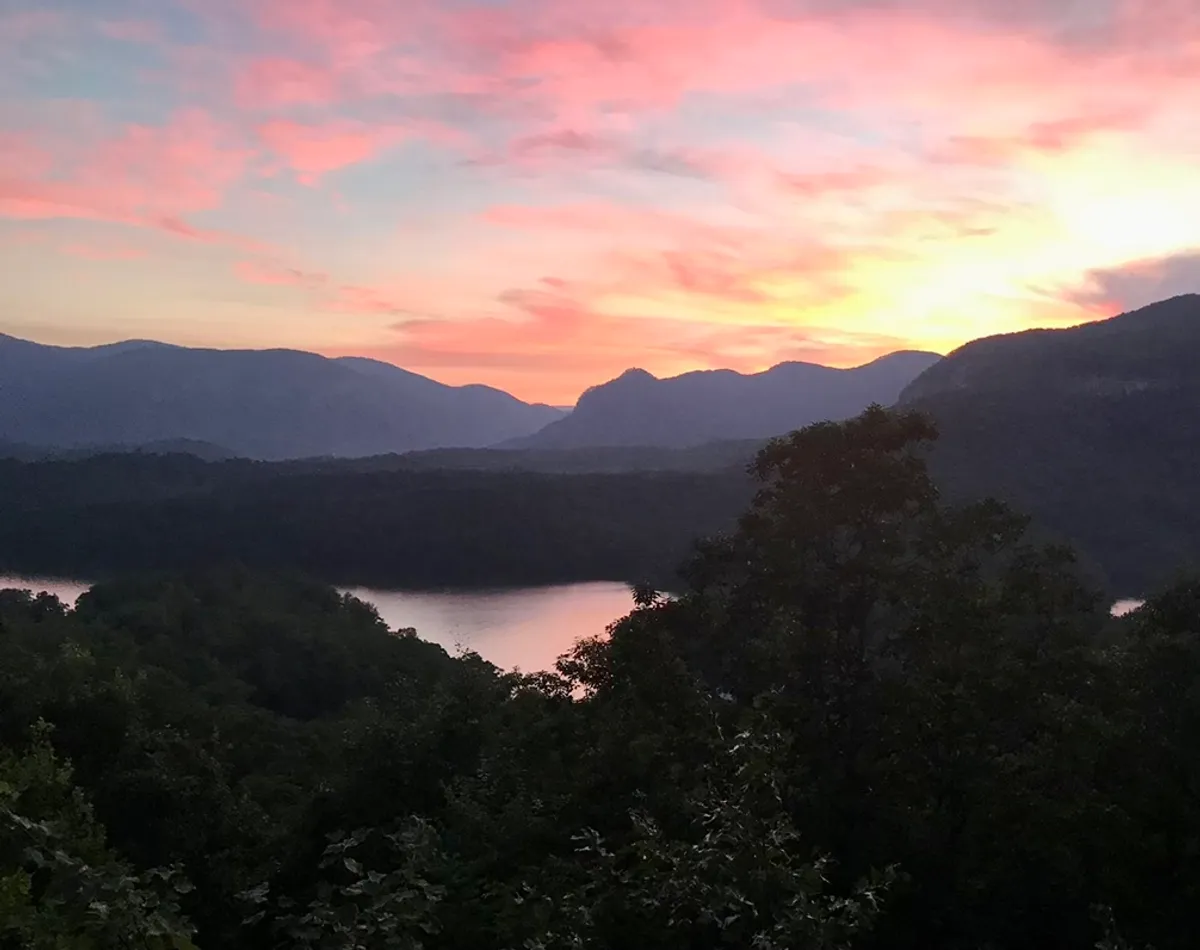 The hero image frames sweeping long-range views over forested mountains and sparkling Lake Lure under a glowing sunset sky, inviting a sense of peace and awe. The vantage point appears to be from a spacious deck or window, emphasizing the cabin’s prime position for soaking in nature's beauty and sharing memorable moments with loved ones.