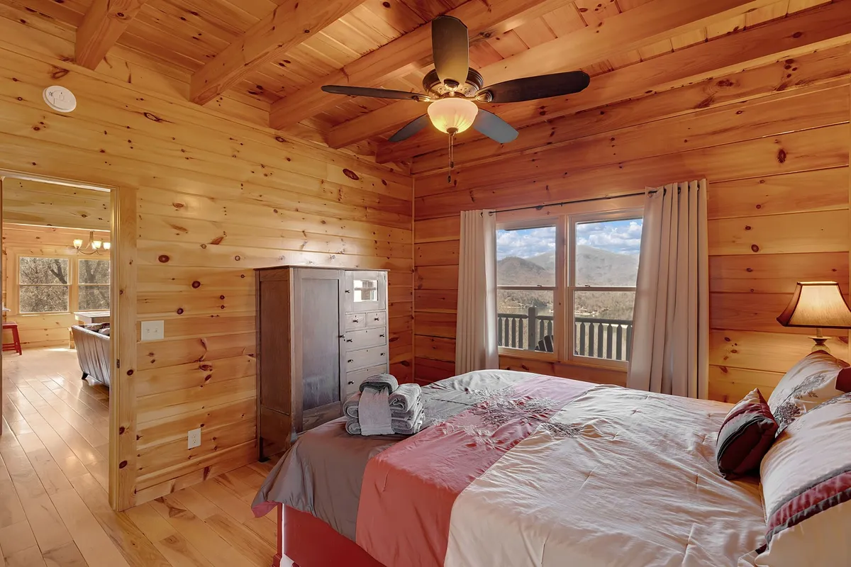 A welcoming bedroom with a large window framing a breathtaking mountain view, a wooden ceiling, a bed adorned with decorative pillows, and a nightstand with a lamp. The setting invites relaxation and quiet moments.